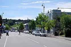 A busy small-town straight street, flanked by shops and the occasional small tree. In the foreground is a pedestrian crossing. Three people are crossing right-to-left – two ladies stepping off the crossing and another entering it. Another lady is crossing left-to-right about halfway across. In the background is a Shell petrol station and behind that a small hill with a large house about halfway up.