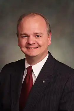 A middle-aged white man is wearing a dark suit, white shirt, and red tie; he has thinning brown hair and is smiling while looking into the camera.