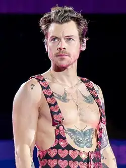 A brown-haired man with light facial hair and a serious expression shows off large bird and butterfly tattoos by being barechested, except for artistic overalls decorated in hearts.