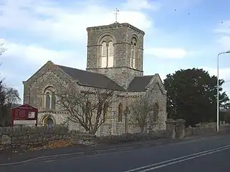 Stone building with arched windows and central square tower. In the foreground are trees and a road.