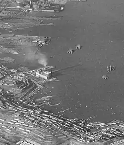 A black and white photograph of the San Francisco Bay shoreline at Hunters Point. The Hunters Point Power Plant is distinguished in the center of this cropped photograph as a large facility emitting smoke from a tall smokestack.