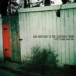 A photograph of a run-down wooden shack with a red door with "DAN ANDRIANO IN THE EMERGENCY ROOM hurricane season" written over it black. The 'M' in "emergency" and the second 'S' in "season" are coloured red
