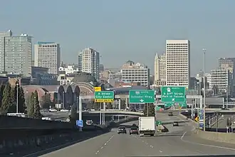 Several high-rise buildings in the background behind a freeway with light traffic and several overhead destination signs