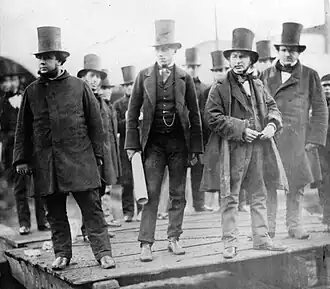 A group of ten men in nineteenth-century dark suits, wearing top hats, observing something behind the camera