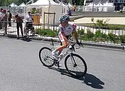 A road racing cyclist in a white jersey with red trim atop a white bicycle, looking behind him out of the frame. Spectators are visible on the roadside behind a barricade.