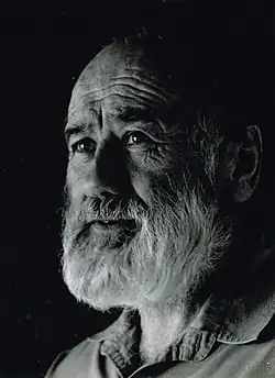 Photographic portrait in black and white. John Bevan Ford is an older man in this photo, with a grey beard and expressive face. He looks off to the side.