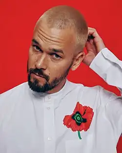 Justin Nozuka wearing a white, collarless shirt with a poppy flower over the left breast pocket, head tilted to the right with left hand scratching back of head, looking directly at camera