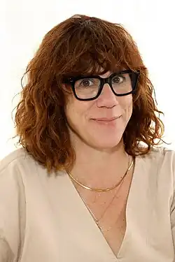 Headshot of Attenberg smiling at the camera. She wears curly hair shoulder-length with bangs and a tan top.