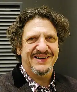 A middle-aged man with a salt-and-pepper goatee and neck-length curly black hair wearing a purple-spotted white shirt and black jacket.
