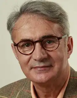 Colour publicity headshot of a half-smiling, clean-shaven, bespectacled, grey-haired man, with strong features