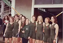 Nine smiling young women, eight in green one-piece tight lycra outfits and the shortest in jumper and trousers, stand in a line in front of two crossed oars