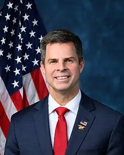 Official House portrait of McGuire smiling in front of the U.S. flag, wearing a black suit with American flag lapel pin with the word "TRUMP" as well as a Navy SEALs badge, white shirt, and red tie.