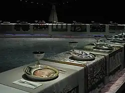 A large, triangular dining table with ornately decorated plates and tableclothes in a dark room. The space encapsulated between the three sides of the table shows a floor with the written names of famous women.