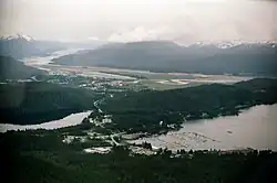 Aerial view shows Auke Bay (including the harbor and Auke Lake) in the foreground. The Mendenhall Peninsula extends to the right behind the community. The lower Mendenhall Valley, Juneau International Airport and Douglas Island are in the background.