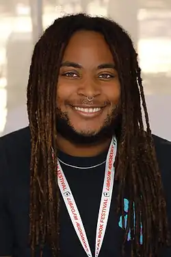 KB Brookins, a Black American writer with brown skin, poses with their hands in their pockets. They are wearing a pink shirt, blue jeans, and a Black belt. The background is purple and green.