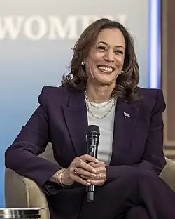 A woman in a purple suit jacket smiles, holding a wireless microphone with both hands while sitting relaxed in a chair.