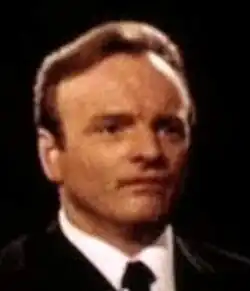 Colour, three-quarter-profile, Press shot of a clean-shaven man with brown hair brushed back, and a serious expression.