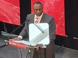 Howell pictured from a above to the left, giving a speech in a black suit at a podium