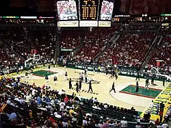 Interior of a basketball arena with a sparse crowd and empty red seats in many sections. A large video board hangs above the court, where players are warming up in small groups.