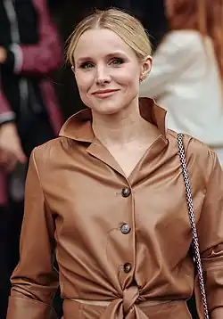 Frontal photo of Kristen Bell from head to waist, wearing a brown leather coat, and with a bag's strap over her left shoulder