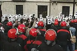 Demonstrators holding pictures in front of uniformed people in red helmets, perhaps police officers