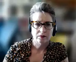 A woman with white hair who is wearing large glasses. The background is blurred because she was appearing via ZOOM.