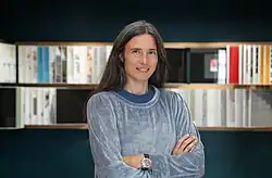 Manuela Pfrunder Smiling, arms crossed, with a soft, blurred background of bookshelves