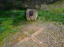 A boulder with a plaque is in the background, with a wooden cross embedded in the ground in the foreground.
