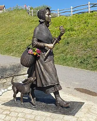 A bronze statue of Mary Anning. She is walking, holding an ammonite with a basket over one arm and her dog trailing behind.