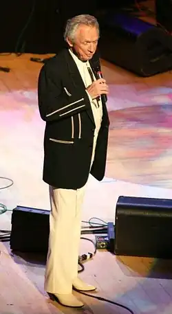 Man in white slacks and black jacket standing singing on stage