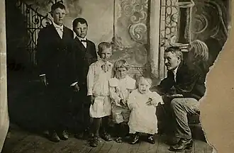 Five children standing in descending order of age and height, adult male sitting next to the youngest one