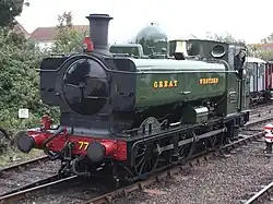 A pannier tank locomotive is passing a station platform, running round its train which includes rolling stock visible in the background. The pannier tanker is green, apart from the black chimney, brass safety valve cover, red buffer beam, and grey coupling rods. Great Western is written on the side of the tank, and 7752 on a number plate on the cab