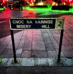 Picture of a street sign on a pavement. The sign is narrow black board with two metal legs embedded on the pavement. It has "Cnoc na hAinnise" written in white. Below that "Misery Hill" is written in white. The right side of the sign is white, with "2" written in white. In the background, there are lights reflecting on the ground.