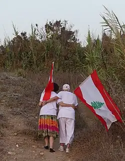 An elderly couple, each holding a flag of Lebanon while walking on the beach
