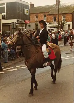 A woman riding a horse in a village centre, surrounded by a crowd.