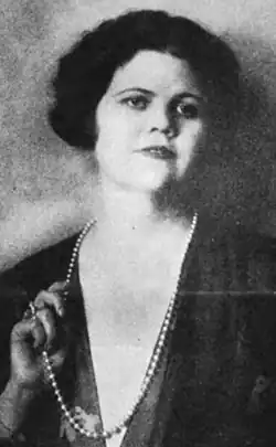 A white woman with dark bobbed hair, wearing a dark translucent dress with a deep-v neckline, and a strand of beads or pearls