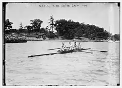 Four men sitting in a long canoe, in front of a wooded camp