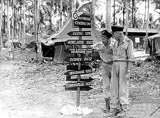 Two men in light-coloured uniforms with a sign reading "Perth 5290, Adelaide 3507, Young & Jacksons Pub Melbourne 2990, Sydney 2472, Brisbane 1955, Townsville 1237"