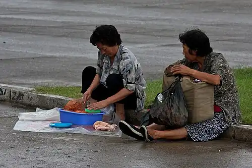 Two elderly North Korean women sitting on a street. One of them is preparing food.