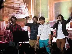 Four teenage boys standing next to each other and singing on a stage. All four are wearing dress shirts and trousers. Left to right: Zayn Malik, Liam Payne, Louis Tomlinson, and Harry Styles.