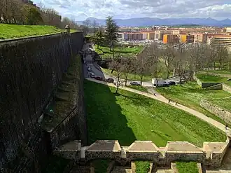 Photo shows an old wall at left and modern buildings at right with mountains in the distance.