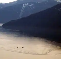 Photograph: top right-hand corner is filled with a shadowed mountain. The lower half shows a small section of Lake Pend Oreille. An object in the bottom right-hand corner is said to be the Paddler.