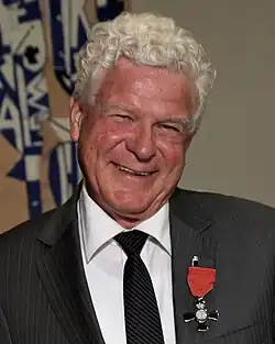 An older man with curly white hair smiles at the camera. He is wearing a dark pinstripe suit, white shirt, and black tie. Pinned to his lapel is a New Zealand honour medal with a red ribbon and silver cross. The background features abstract blue and beige artwork.