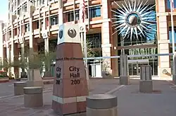 photos showing the short obelisk signage showing City Hall, and topped with the seal of the city, a stylized maroon phoenix. The semi-circular front of the building in the background, adorned with a stylized sunburst.