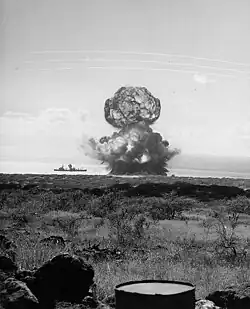 Shot Bravo, the first test, with the test ship Atlanta (IX-304) moored nearby. Note smoke or dust around the ship's foremast, and the shock wave perimeter expanding on the water beyond the ship