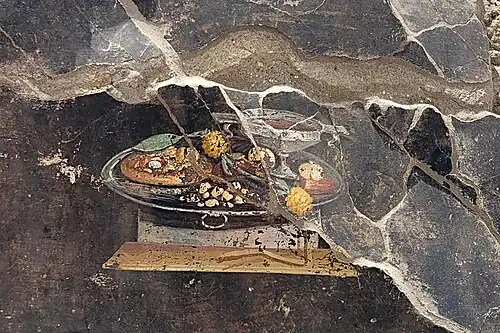 Fresco in the hallway depicting a goblet of wine and various fruits, including a pomegranate and date. The pizza-like bread appears to have moretum, a cheese spread with herbs popular in ancient Rome. The fresco is likely to be inspired by the Greek hospitality ritual of xenia and shows gifts typically offered to guests[12]