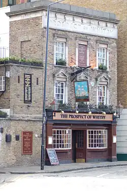 A narrow, brick pub with white-framed windows, a dark entrance, and hanging signs, viewed from a cobbled street.