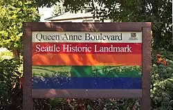 photograph of a sign created by the Parks and Recreation department for Queen Anne Boulevard. It has horizontal stripes in cream, red, orange, green, and blue (from top to bottom)