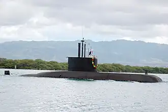 A submarine travelling on the water's surface near to shore, with trees and mountains in the background.