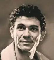 Sepia publicity headshot of a smiling, clean-shaven, lean man with black curly hair and strong features